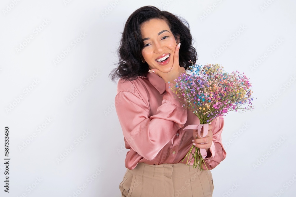 young woman holds big bouquet of nice flowers over white background looking confident at the camera smiling with crossed arms and hand raised on chin. Thinking positive. young woman holds big bouquet of nice flowers over white background looking confident at the camera smiling with crossed arms and hand raised on chin. Thinking positive.