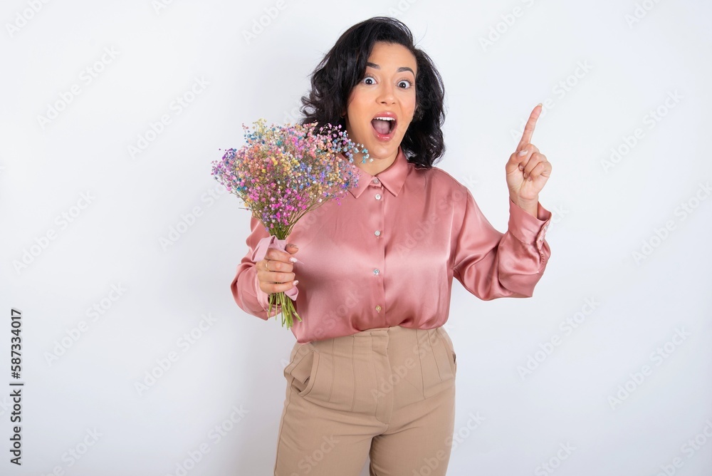 young woman holds big bouquet of nice flowers over white background pointing finger up and looking inspired by genius thought, showing good idea sign, having clever solution in mind young woman holds big bouquet of nice flowers over white background pointing finger up and looking inspired by genius thought, showing good idea sign, having clever solution in mind