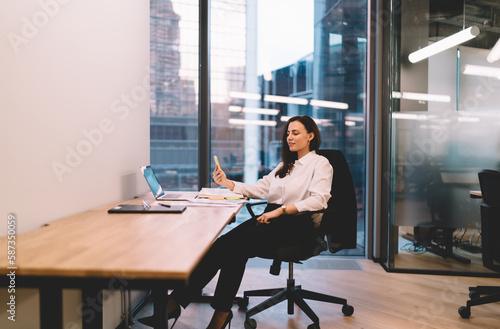 Woman with smartphone in hand sitting with laptop in office