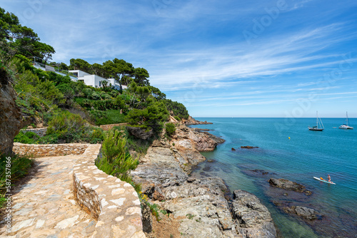 Coastal path along the cliff with view to sea, anchored yachts and padling couple - June 2018 - Sa Riera, Costa Brava, Catalonia, Spain