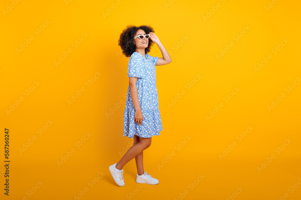 Full length photo of pretty, fashionable curly brazilian or african american woman in a blue summer dress and white sneakers, with trendy sunglasses, looking away, smiling, isolated orange background Full length photo of pretty, fashionable curly brazilian or african american woman in a blue summer dress and white sneakers, with trendy sunglasses, looking away, smiling, isolated orange background