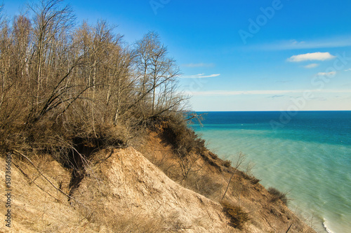 Fototapeta Naklejka Na Ścianę i Meble -  Under a sunny blue sky in early Spring, the view of a peaceful Lake Michigan as seen from atop a sandy, forested bluff at Lion's Den Gorge, near Grafton, WI.