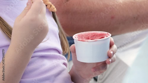 Anonymous elementary school age child girl eating ice cream from a small white blank paper cup with a spoon, closeup, detail, one person. Kids eating sweet food, nutrition, sugar simple concept