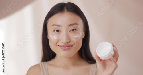 Portrait of Asian woman applying skin care cream on face. Cute female smiling and happy, using product for young skin. Moisturizing and having shining skin. Dermatology, cosmetology concept. photo