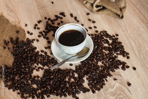 A cup of hot coffee in a plate with a spoon. Coffee beans are scattered on a wooden light brown table.