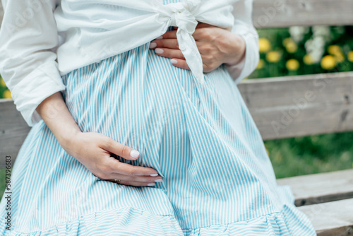 A pregnant woman strokes her stomach in the park on a bench, hands on her stomach close-up.Pregnancy, women's health concept.