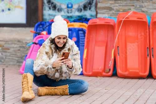 Smiling middle-aged woman dressed in warm clothing using her smartphone in front of a sled rental shop