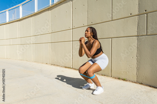 young black girl doing squat exercise with elastic band in sportswear. © Dani