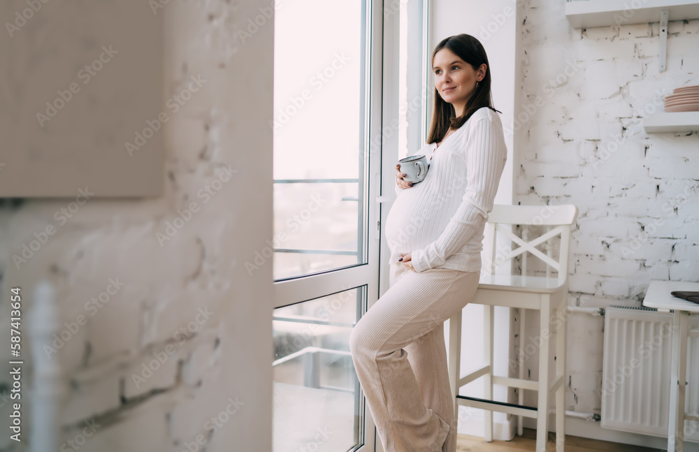 Fototapeta premium Smiling pregnant woman in white outfit leaning on high raised chair at home
