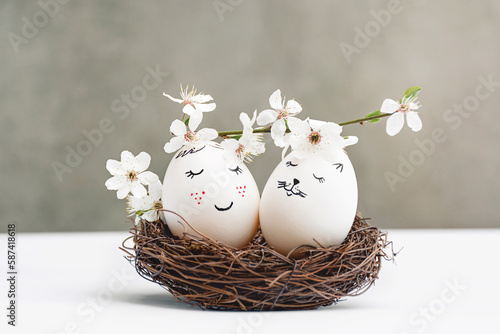 Happy Easter greeting card. White Easter eggs in wicker wooden nests on white table on gray background. Gentle painted faces. View is direct. Blossoming branch of cherry with white flowers. Copy Space
