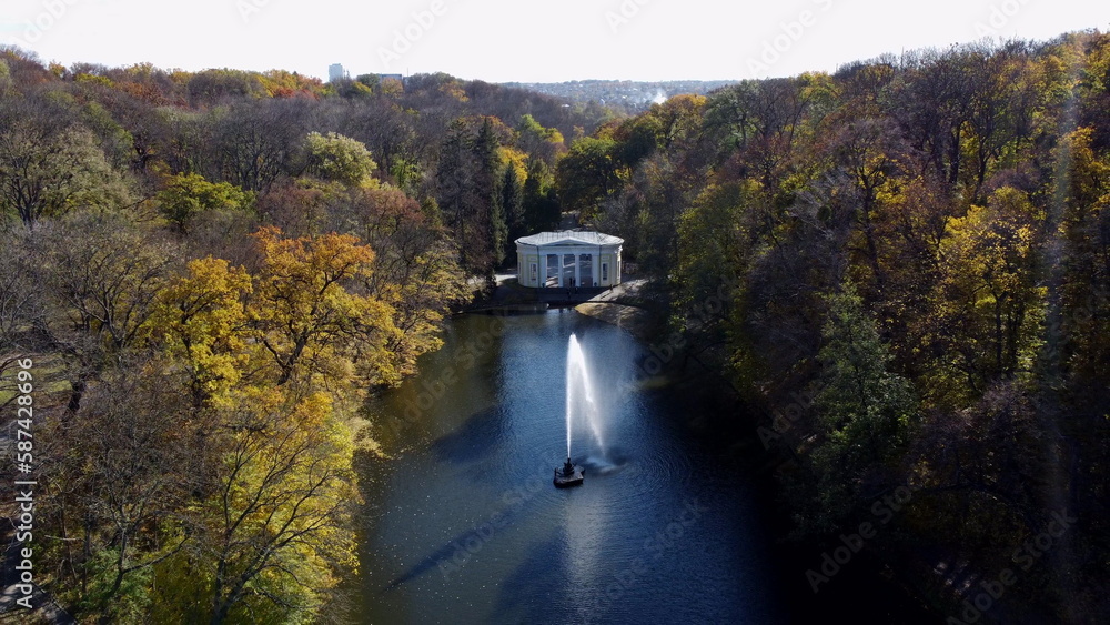 Beautiful landscaped park with lake, fountain, trees, large stones, people walking and white architecture on sunny autumn day. Trees with yellow leaves on shores, tourists high flowing jet of fountain Beautiful landscaped park with lake, fountain, trees, large stones, people walking and white architecture on sunny autumn day. Trees with yellow leaves on shores, tourists high flowing jet of fountain