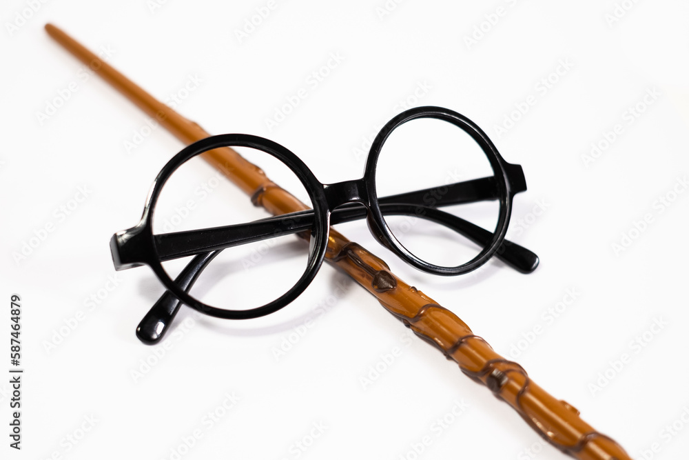 Fototapeta premium Round black eyeglasses and brown magic wand isolated on white background. Harry Potter style circle glasses and wand. Wizard costume accessories. 