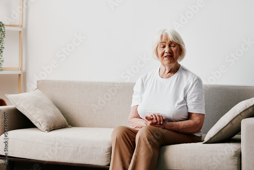 Elderly woman sits on sofa at home, bright spacious interior in old age smile, lifestyle. Grandmother with gray hair in a white T-shirt and beige trousers.