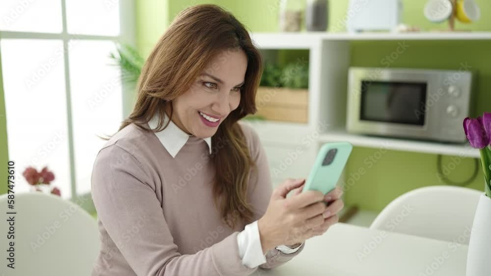 Young beautiful hispanic woman using smartphone sitting on table at home Young beautiful hispanic woman using smartphone sitting on table at home