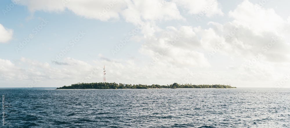 From the vantage point of a boat in the blue sea, this captivating shot of an island with lush greenery and trees. White clouds dot the sky, and a single antenna stands tall against the horizon