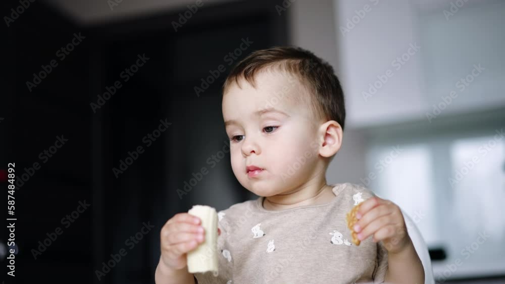 Little kid is eating holding food in both hands. Lovely baby boy eating banana. Low angle view. Little kid is eating holding food in both hands. Lovely baby boy eating banana. Low angle view.