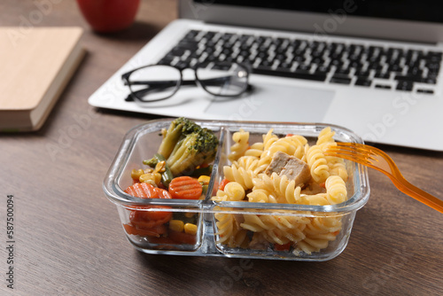 Container of tasty food, fork, laptop and glasses on wooden table. Business lunch