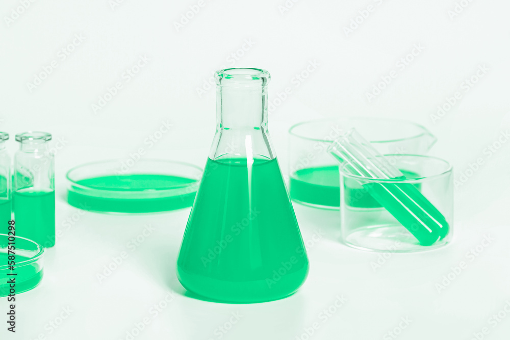 Flask for a laboratory with a green liquid. Against the background of laboratory glassware. Petri dishes, flasks, test tubes. On a white, light background. Flask for a laboratory with a green liquid. Against the background of laboratory glassware. Petri dishes, flasks, test tubes. On a white, light background.
