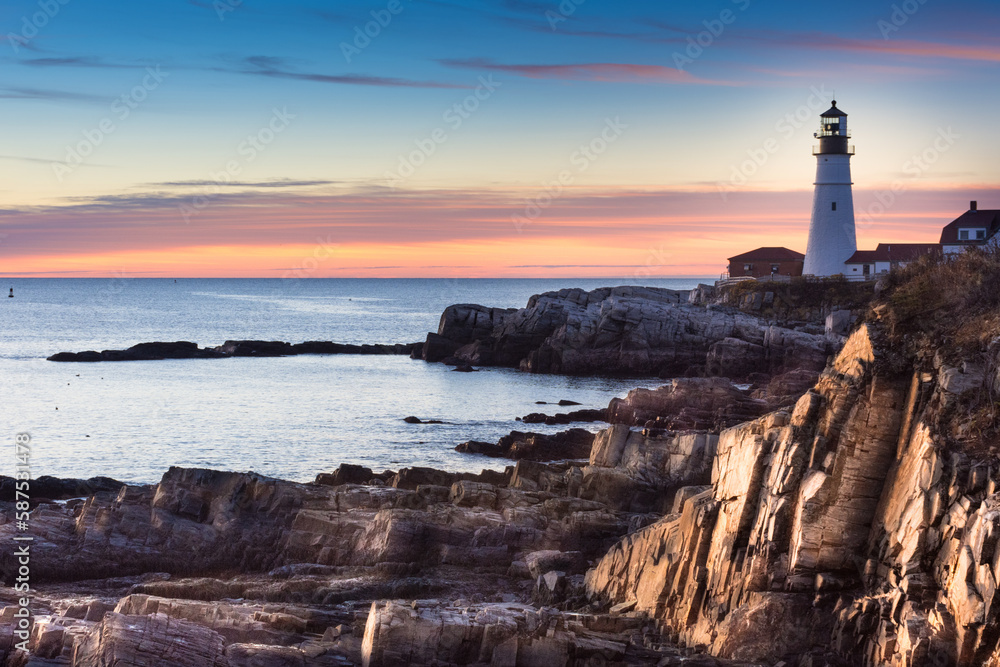 Sunrise at Portland Head Lighthouse, Portland, Maine. Shot from the north looking across cliffs bathed in golden sunlight. Clouds and sky at horizon fiery orange with blue sky above.