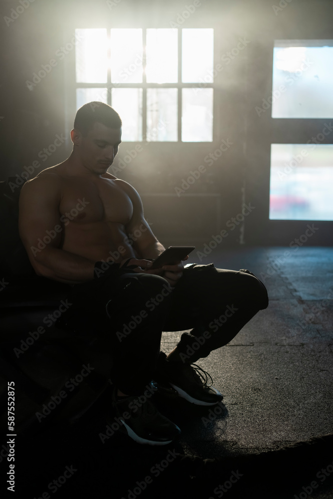 Shirtless male athlete with six pack abs using tablet while sitting on a large tyre in sports gym Shirtless male athlete with six pack abs using tablet while sitting on a large tyre in sports gym