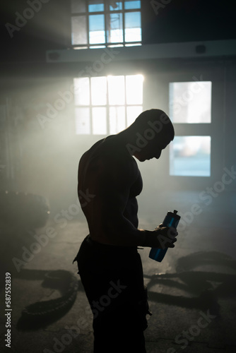Shirtless male athlete drinking water from plastic bottle after workout inside abandoned warehouse