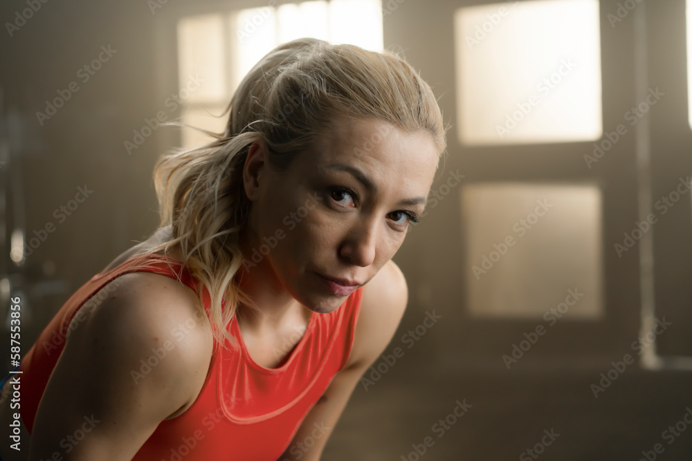 Fototapeta premium Tired athlete leans forward and looks at the camera during a rest break in a gym. Close-up fitness portrait of a woman in a red training top, with warm window light behind her.