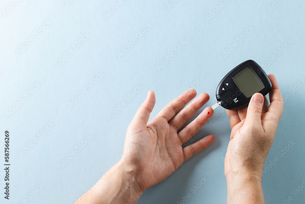 Fototapeta premium Hands of caucasian woman taking blood sugar reading with glucometer, on blue background