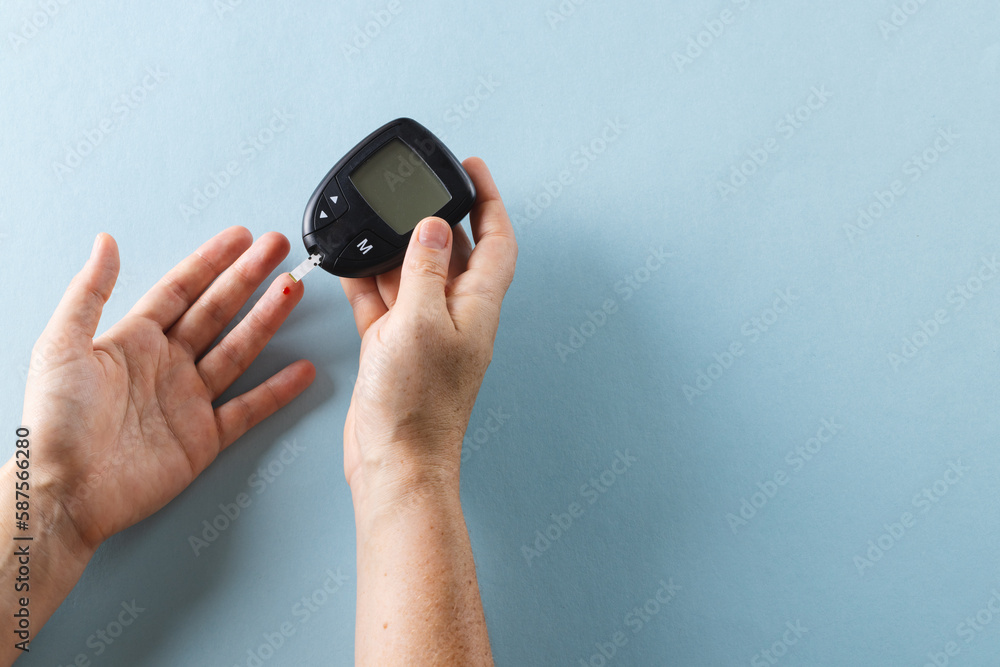 Fototapeta premium Hands of caucasian woman taking blood sugar reading with glucometer, on blue background, copy space