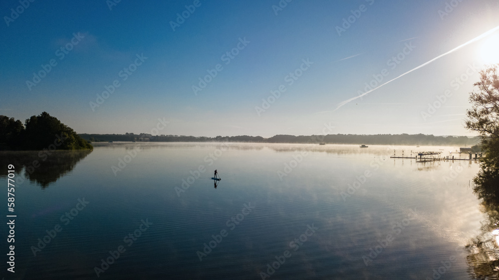 Woman paddling on SUP board on beautiful lake aerial drone view with reflections from above. Standing up paddle boarding adventure in early morning sunrise. Germany lake district Mecklenburg.
