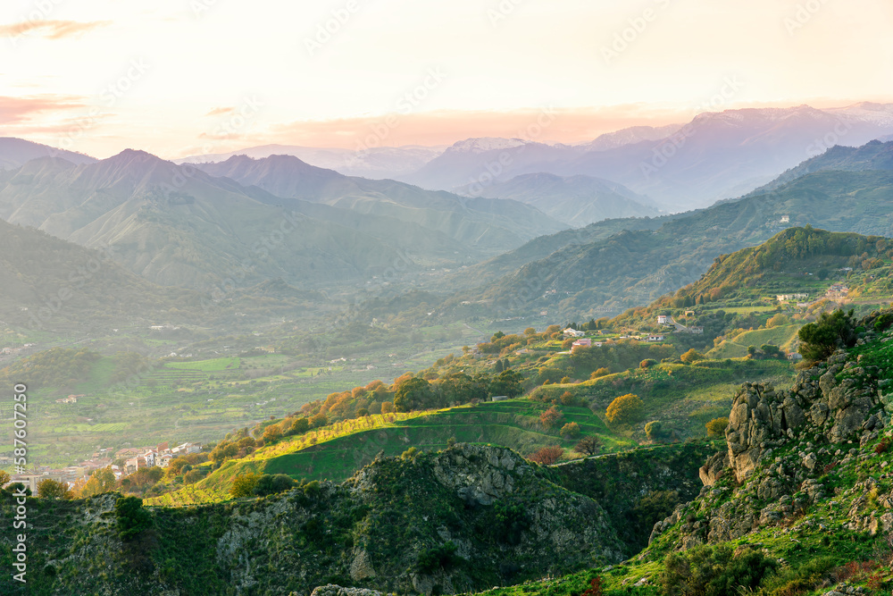 picturesque view in a green mountain valley among slopes and hills with sunny plantations on a rocky plato and nice cloudy sunset on background