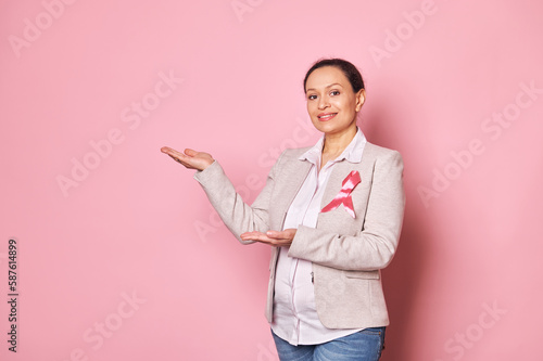 Pleasant pregnant woman wearing pink satin ribbon - emblem of fighting Breast Cancer, showing a copy advertising space on isolated background. 1st October. Pink month. Breast Cancer Awareness campaign