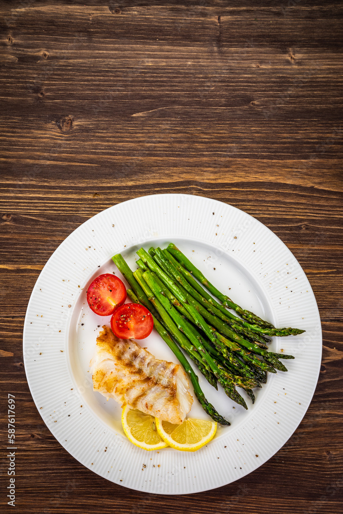 Fish dish - fried cod with green asparagus, lemon and fresh tomatoes on wooden table
