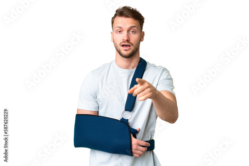 Young caucasian man with broken arm and wearing a sling over isolated chroma key background surprised and pointing front