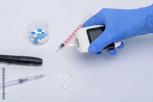 Trzymany w dłoniach w niebieskich rękawiczkach glukometr, obok strzykawki do insuliny, lekarstw
