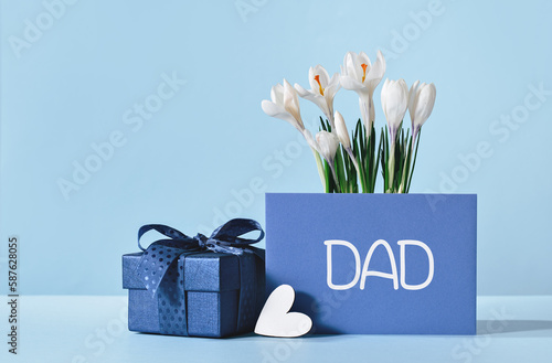 Fototapeta Naklejka Na Ścianę i Meble -  Small luxury gift box with blue bow and white heart. Dad text on gift card and white crocus flowers. Fathers day or birthday gift for him. Festive sale