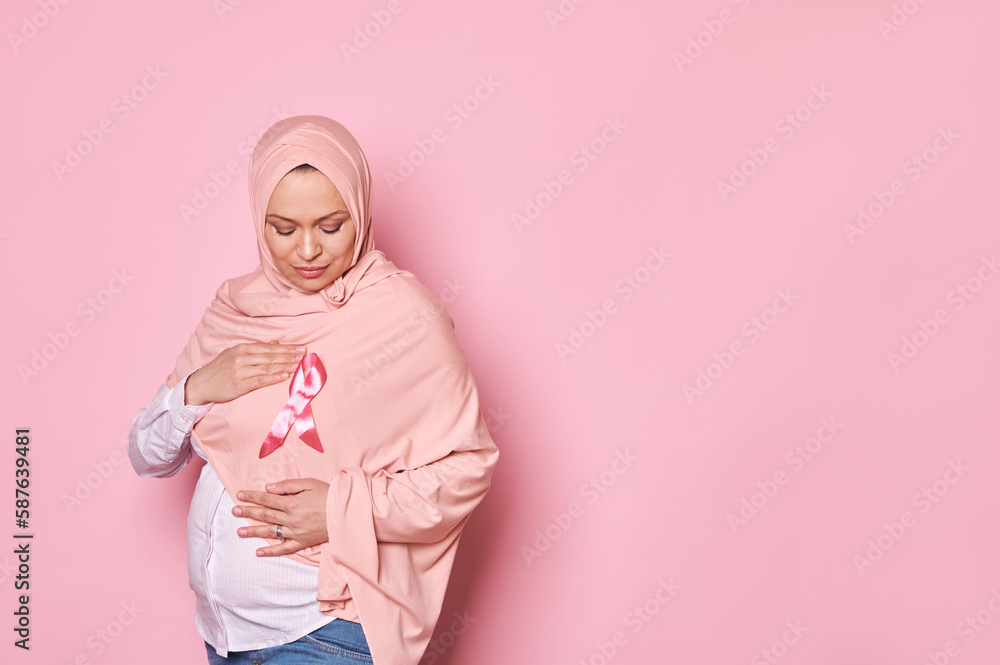 Fototapeta premium Muslim pregnant woman, holds her hands around pink satin awareness ribbon on hijab, shows her support for female survivors and patients. October 1 st - International Day fighting against Breast cancer