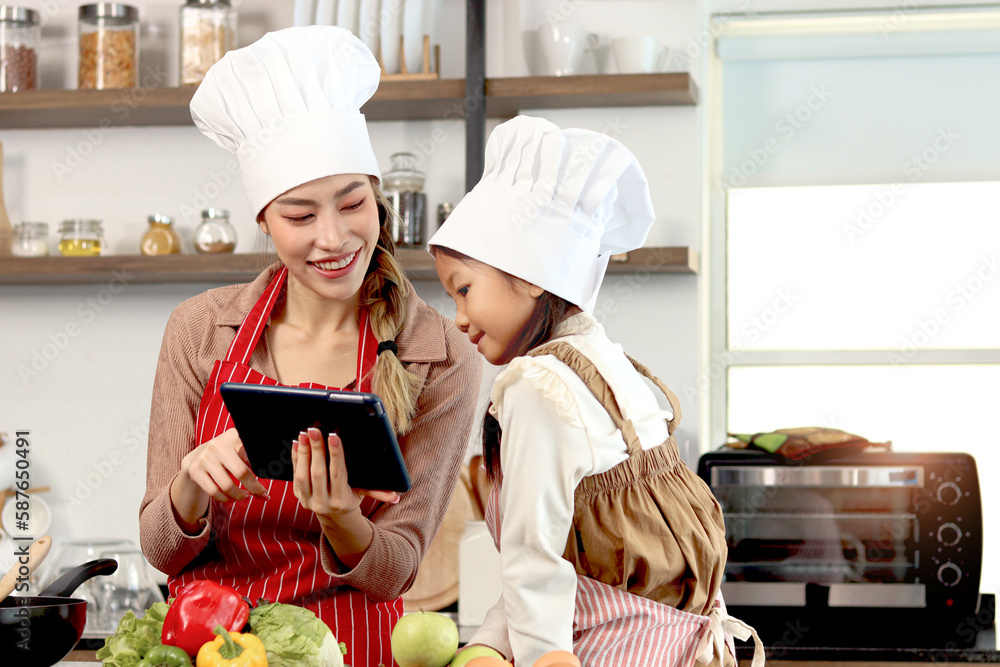 Happy Asian mother daughter wearing apron and chef hat at kitchen, mom and girl kid look at digital tablet for searching tasty healthy meal recipes on internet, cute chef family cooking food at home. Happy Asian mother daughter wearing apron and chef hat at kitchen, mom and girl kid look at digital tablet for searching tasty healthy meal recipes on internet, cute chef family cooking food at home.
