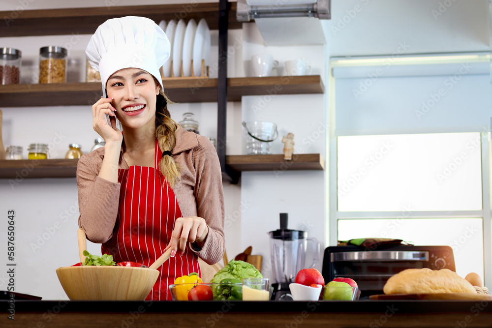 Happy smiling Asian beautiful woman wear apron and chef hat, talking on mobile phone during prepare fresh vegetables salad at kitchen, cute female chef cooking healthy food, vegan salad, diet, dieting Happy smiling Asian beautiful woman wear apron and chef hat, talking on mobile phone during prepare fresh vegetables salad at kitchen, cute female chef cooking healthy food, vegan salad, diet, dieting