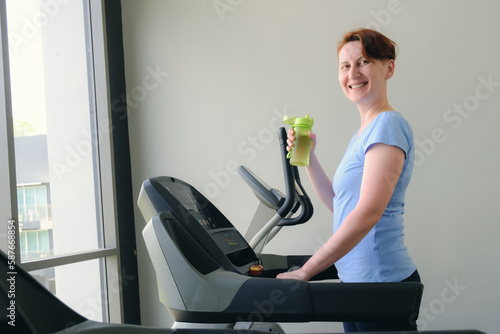 Woman stands on a treadmill in the gym, hold sports bottle with water and smiles. Middle-aged woman in a blue t-shirt goes in for sports. The concept of a healthy lifestyle, weight loss.