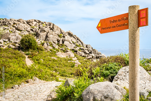 A signpost directs hikers to PR 3 Trilho da Fóia and PR 5 Percurso das Cascatas trails, leading to the sunlit Fóia mountain top with the distant haze-wrapped city of Portimão visible in the background photo