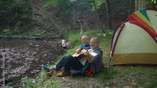 Two gay men enjoying nature camping with tent with pride flag in forest on river playing guitar.