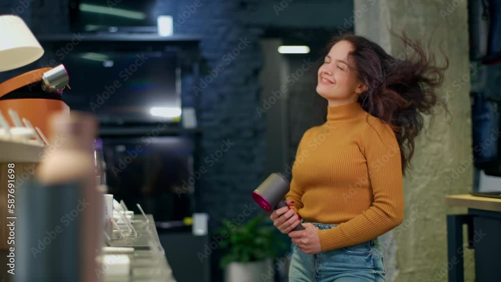 Attractive young girl with curly hair in an orange sweater is testing a hair dryer in a store. The girl holds a hairdryer in her hands, blows hot air through her hair, smiles and looks at the camera Attractive young girl with curly hair in an orange sweater is testing a hair dryer in a store. The girl holds a hairdryer in her hands, blows hot air through her hair, smiles and looks at the camera