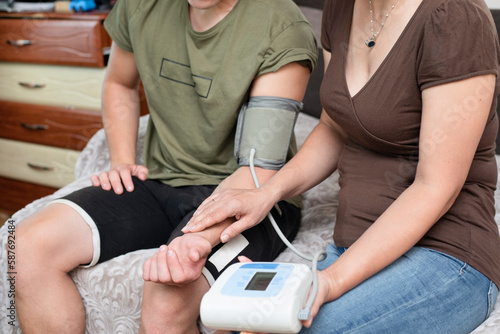 A woman measures her adult son's blood pressure with a torometer and measures pulse with hand, the boy wants to know his physical indicators after training in order to train usefully and safely photo