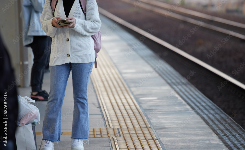 駅のホームで電車を待ちながらスマートフォンを弄る若い女性の姿 駅のホームで電車を待ちながらスマートフォンを弄る若い女性の姿