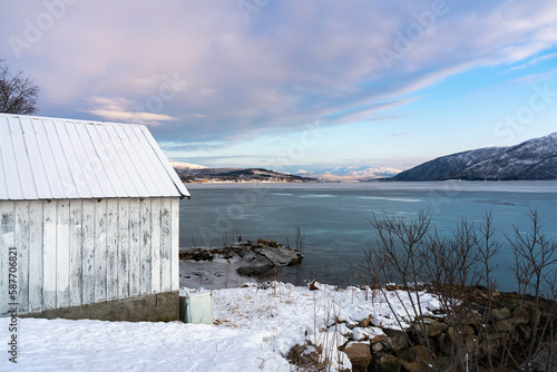 Bootshaus am Ufer des Stonesbotn, Morgenstimmung im Winter mit Eis am Strand des Fjord auf Senja, Norwegen, Morgenrot in den Wolken über dem Meer  photo