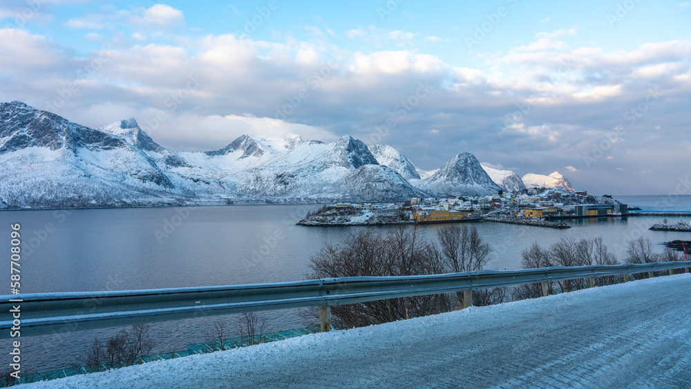 Panorama mit einer Insel mit einem Dorf im Fjord und den schneebedeckten Bergen von Norwegen. Fischerdorf Husøy auf Senja am Morgen im Winter