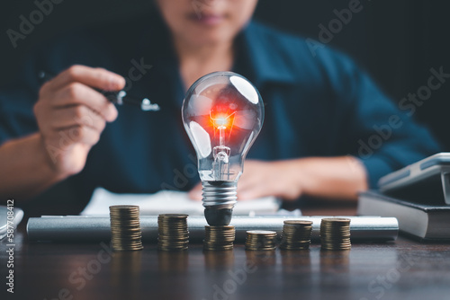 Businesswoman hand holding lightbulb with using calculator to calculate and money stack. Idea of energy saving planning in home. Save energy and money with accounting finance in office concept.