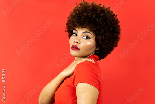 A dark-skinned girl with afro hair poses on a red background. The girl touches her shoulder while looking to the side. African fashion concept for girls with afro hair.