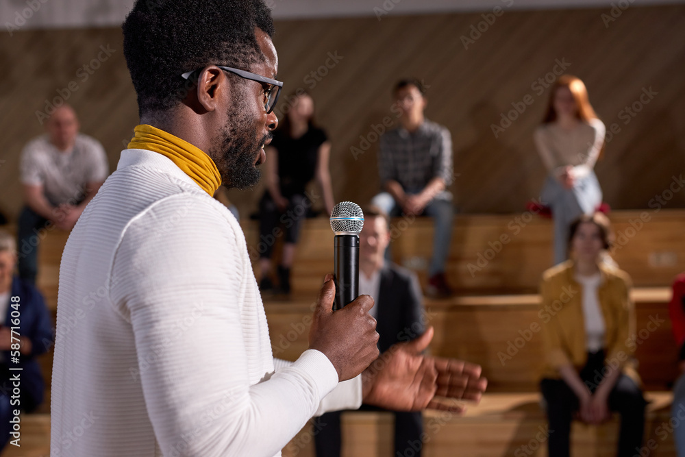 © Roman - African lecturer presenting lesson with microphone to caucasian group of people. Young black teacher talking about finance in lecture hall. Side view on handsome guy in spectacles and white clothing © Roman - African lecturer presenting lesson with microphone to caucasian group of people. Young black teacher talking about finance in lecture hall. Side view on handsome guy in spectacles and white clothing