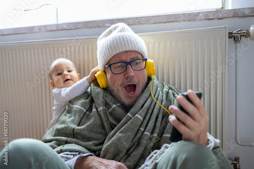 Emotional grey haired matured man in winter hat is sitting near heating at home